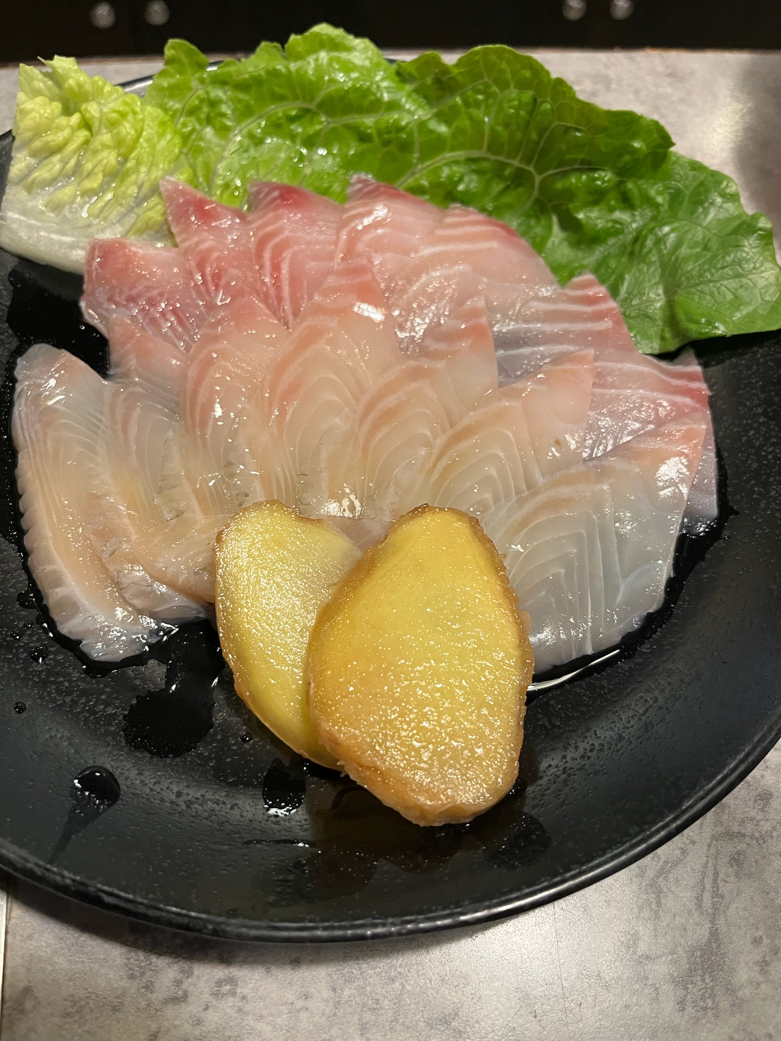 Fresh white fish slices on a black plate, served with pickled ginger and lettuce